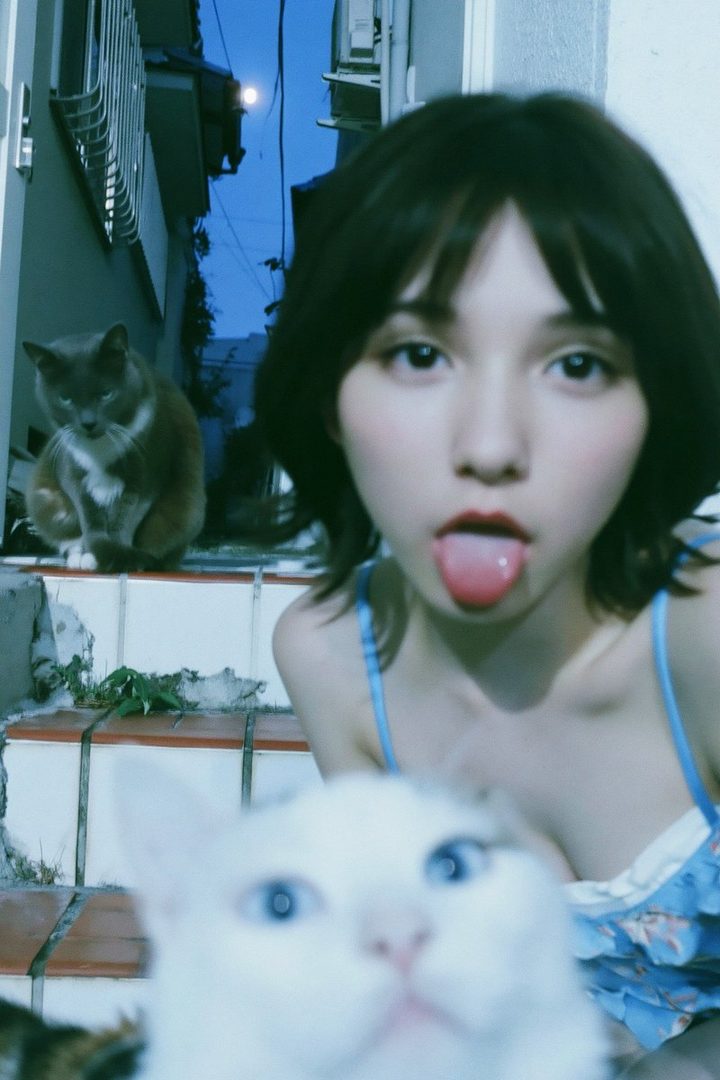 A young woman with short black hair sticks her tongue out playfully at the camera, with two cats in the foreground and background, outdoors at night.
