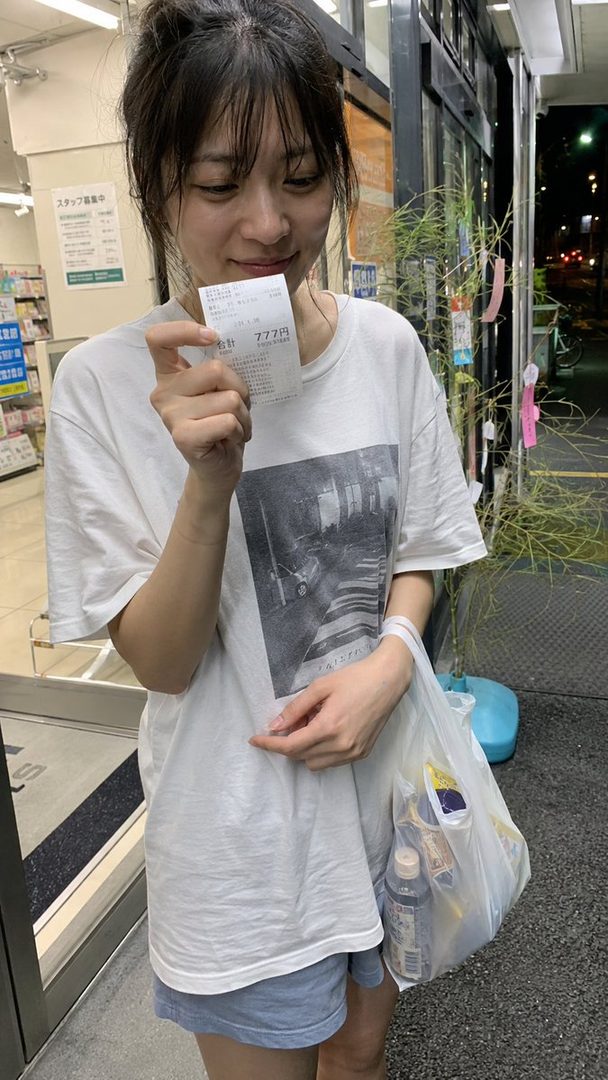 A young East Asian woman in a white t-shirt and shorts holds up a receipt and a shopping bag, smiling slightly.