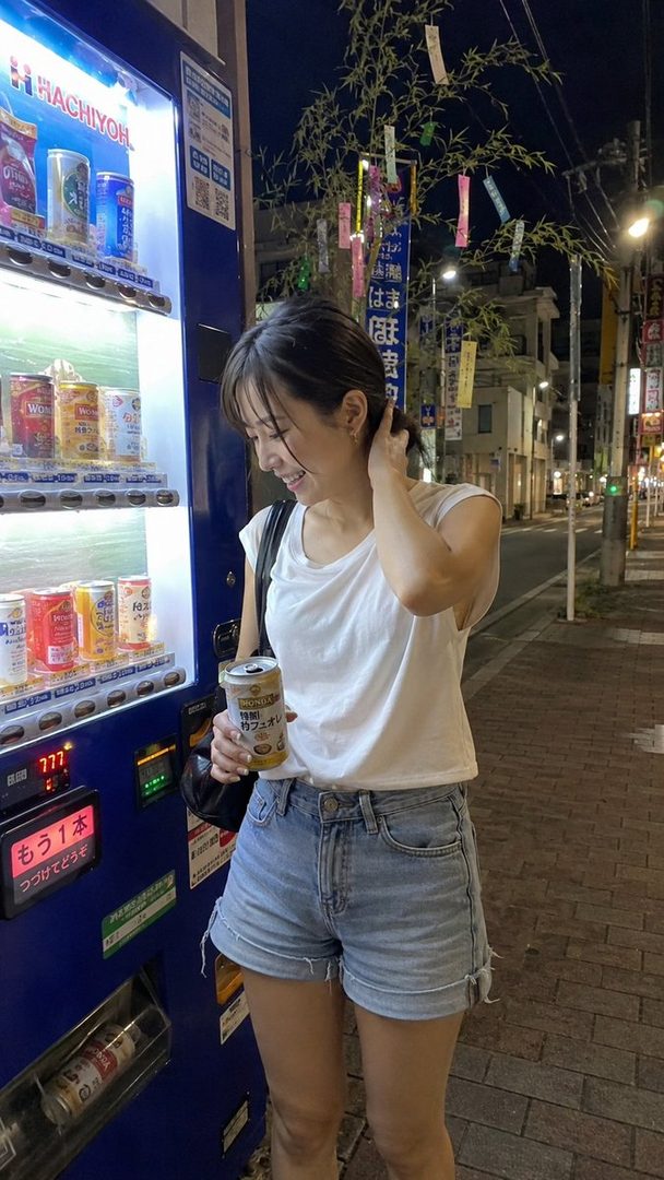 A young Japanese woman in a white t-shirt and denim shorts stands by a brightly lit vending machine at night, holding a can of drink and smiling.