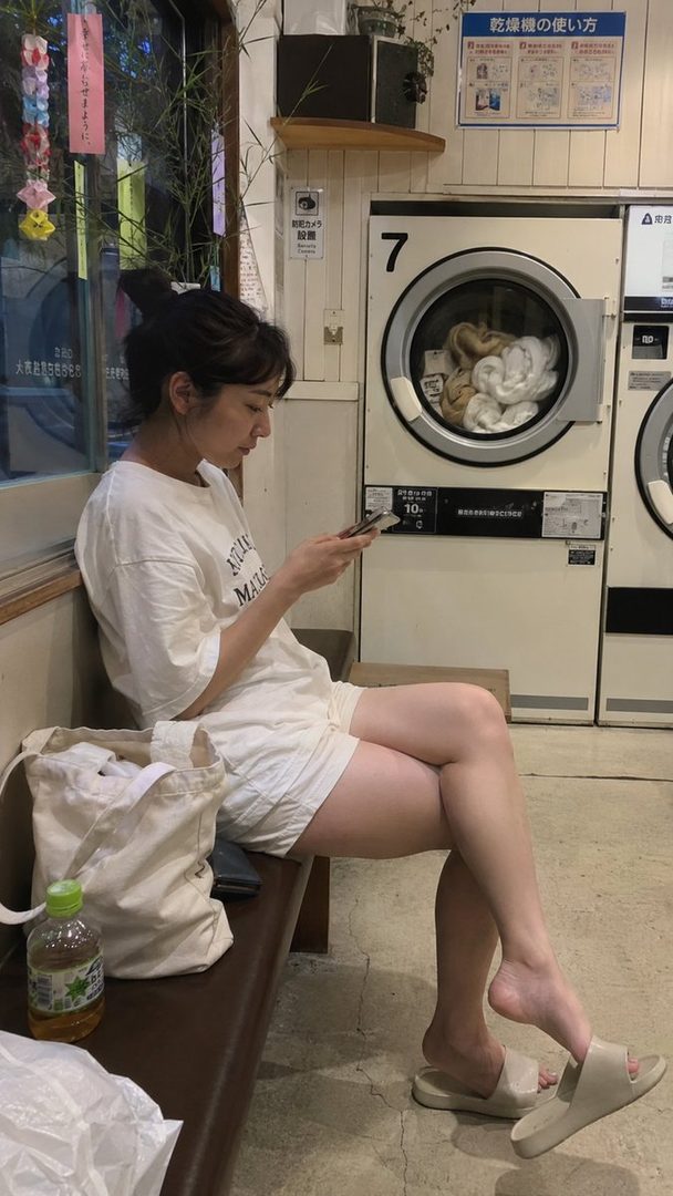 A young woman with her hair in a bun sits in a laundromat, looking at her phone while wearing a white t-shirt and shorts.
