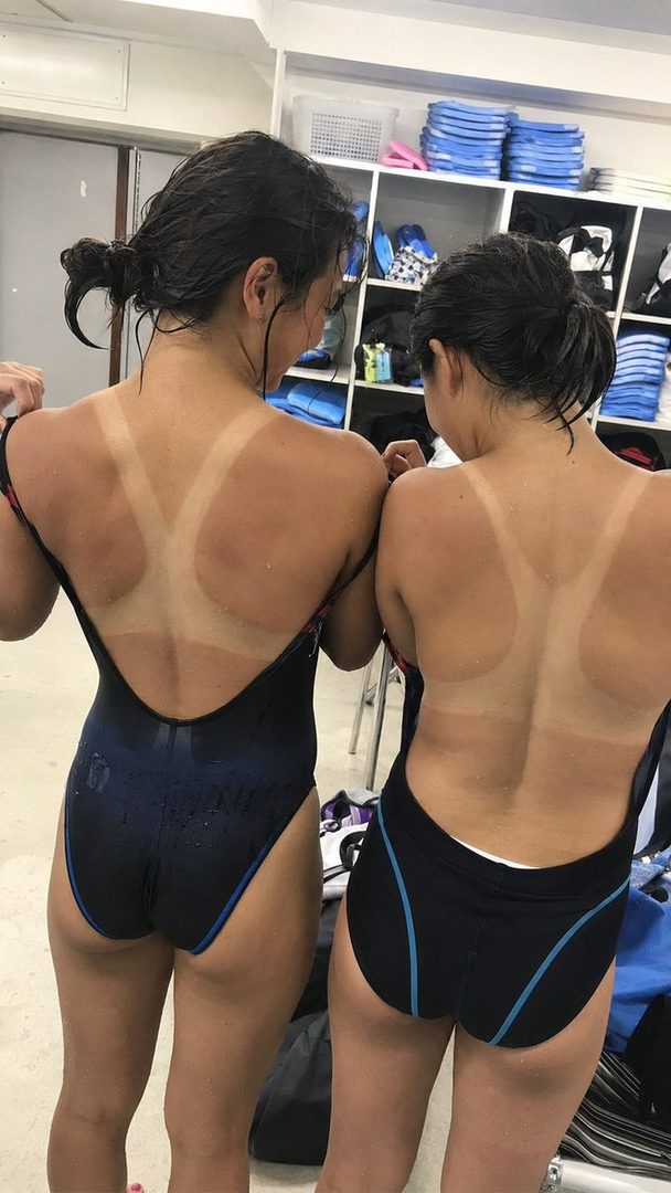 Two young women in swimsuits showing prominent tan lines on their backs, standing indoors in what appears to be a locker room.