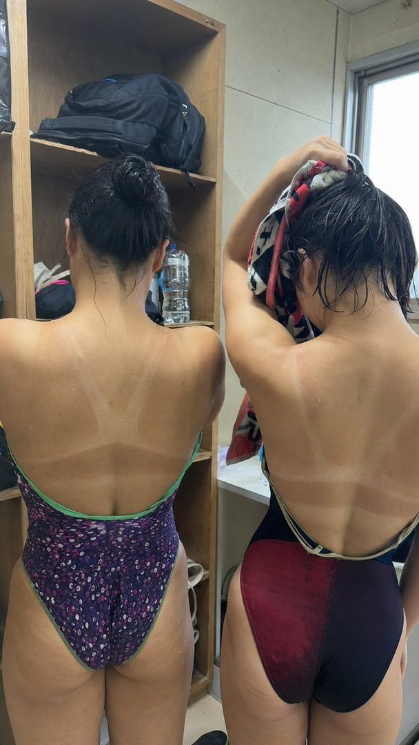 Two women in swimsuits with prominent tan lines are seen from behind in an indoor setting, one drying her hair with a towel.