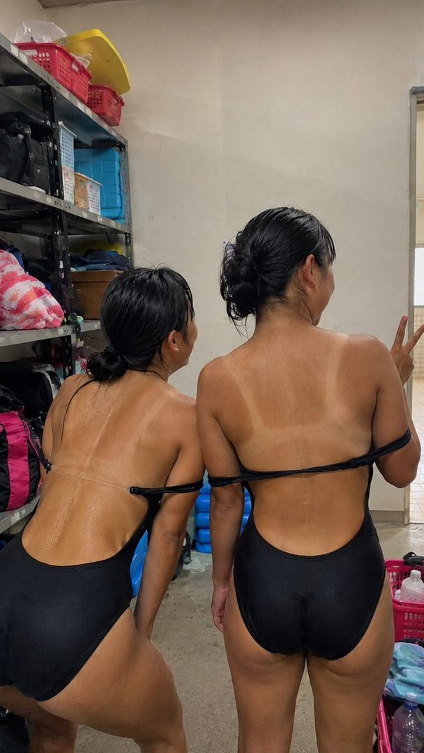 Two women in black swimsuits showing prominent tan lines, posing indoors.