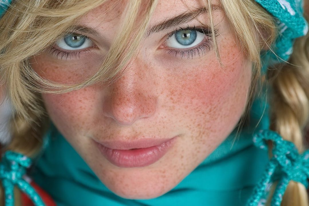 Close-up portrait of a young woman with blonde hair, blue eyes, and freckles, wearing a teal scarf.