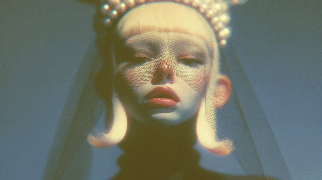A close-up portrait of an AI-generated model with blonde hair, wearing a pearl-adorned headdress and a veil, with dramatic makeup and styling.