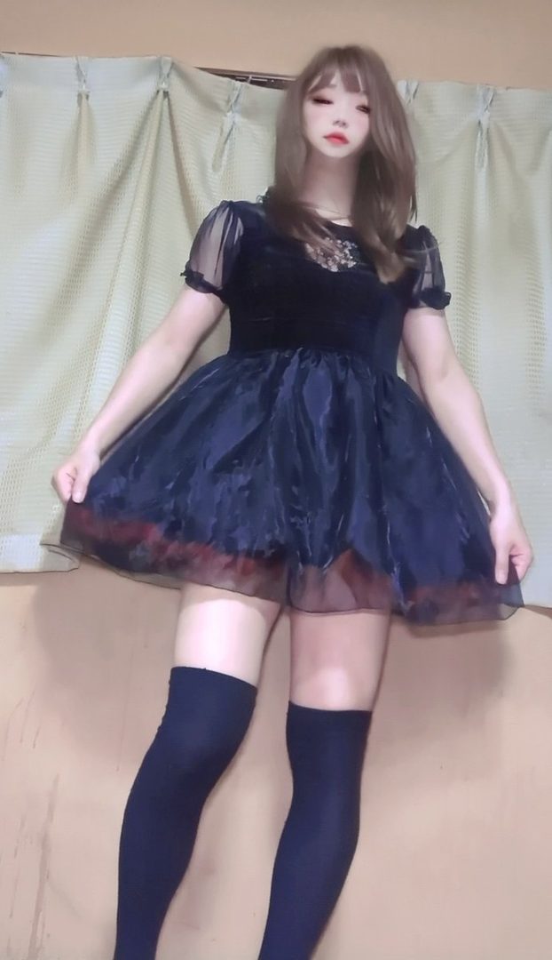 A young woman with long brown hair, wearing a black gothic lolita dress and thigh-high socks, poses indoors.