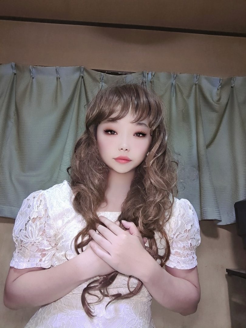 A young woman with long, curly brown hair and bangs, wearing a white lace dress, poses with her hands crossed over her chest indoors.