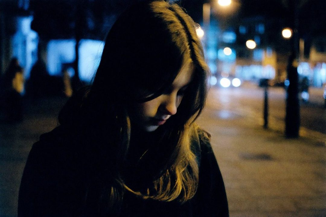 A young woman with long brown hair looks down pensively on a dimly lit city street at night, with blurred lights in the background.
