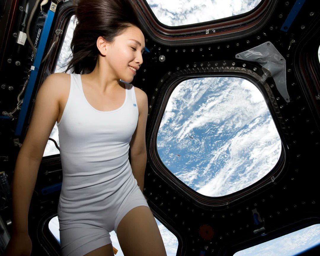 A young East Asian woman in a white tank top and shorts floats inside a spacecraft, smiling as she looks out the window at the Earth below.