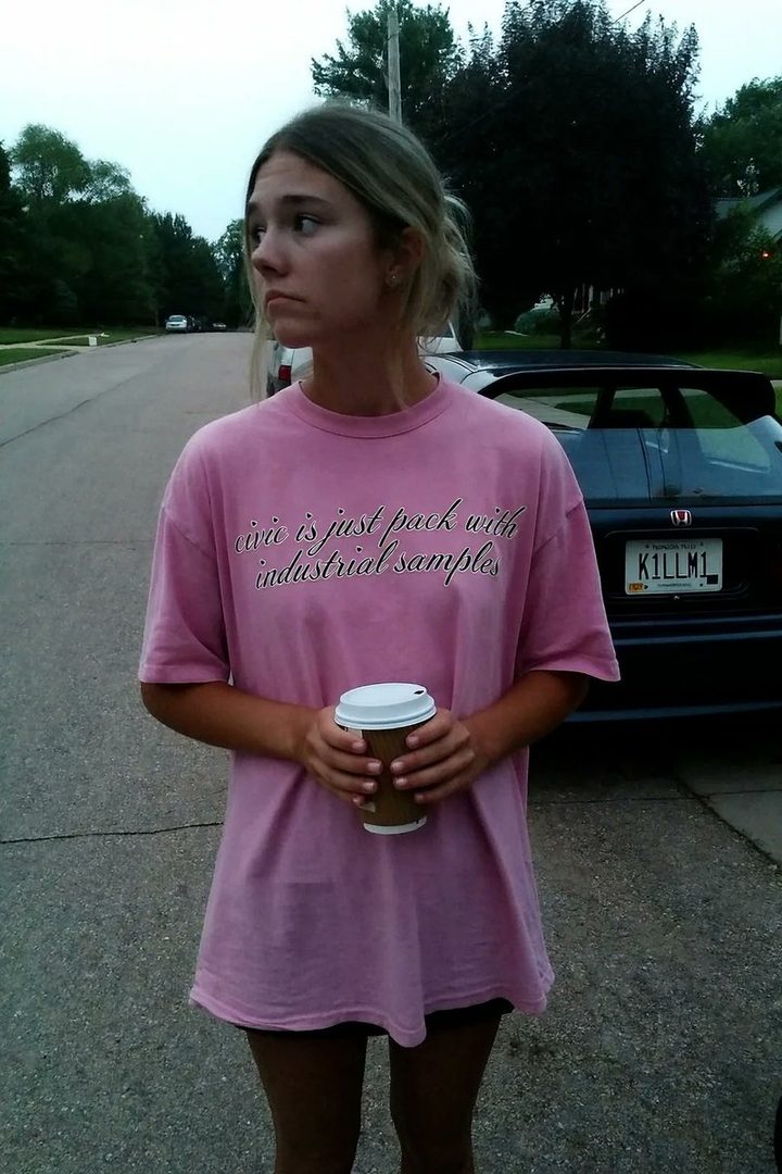 A young woman with blonde hair tied back, wearing a pink t-shirt and holding a coffee cup, stands outdoors on a street.