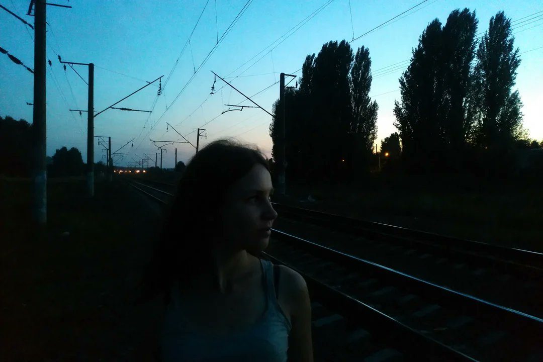 A young woman stands by train tracks at dusk, looking into the distance.