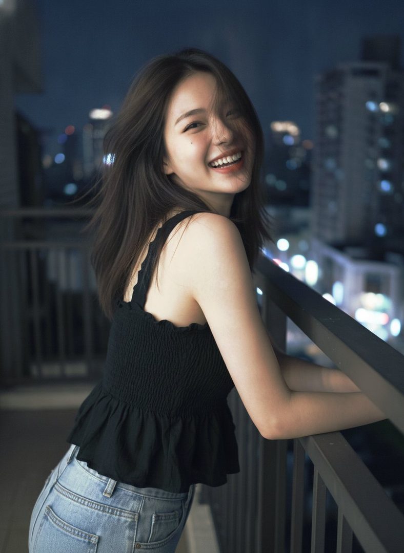 A young woman with long brown hair smiles brightly while leaning on a balcony railing at night, with city lights in the background.