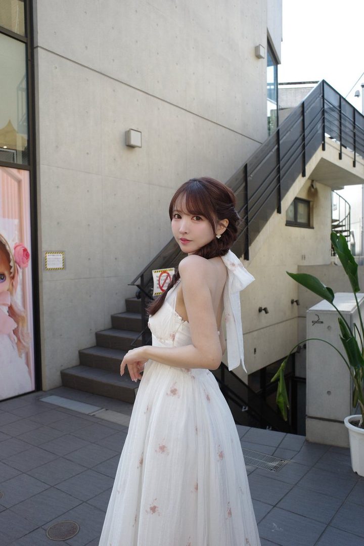 A young woman with brown hair in a braid, wearing a white floral dress, poses outdoors with a modern building and stairs in the background.