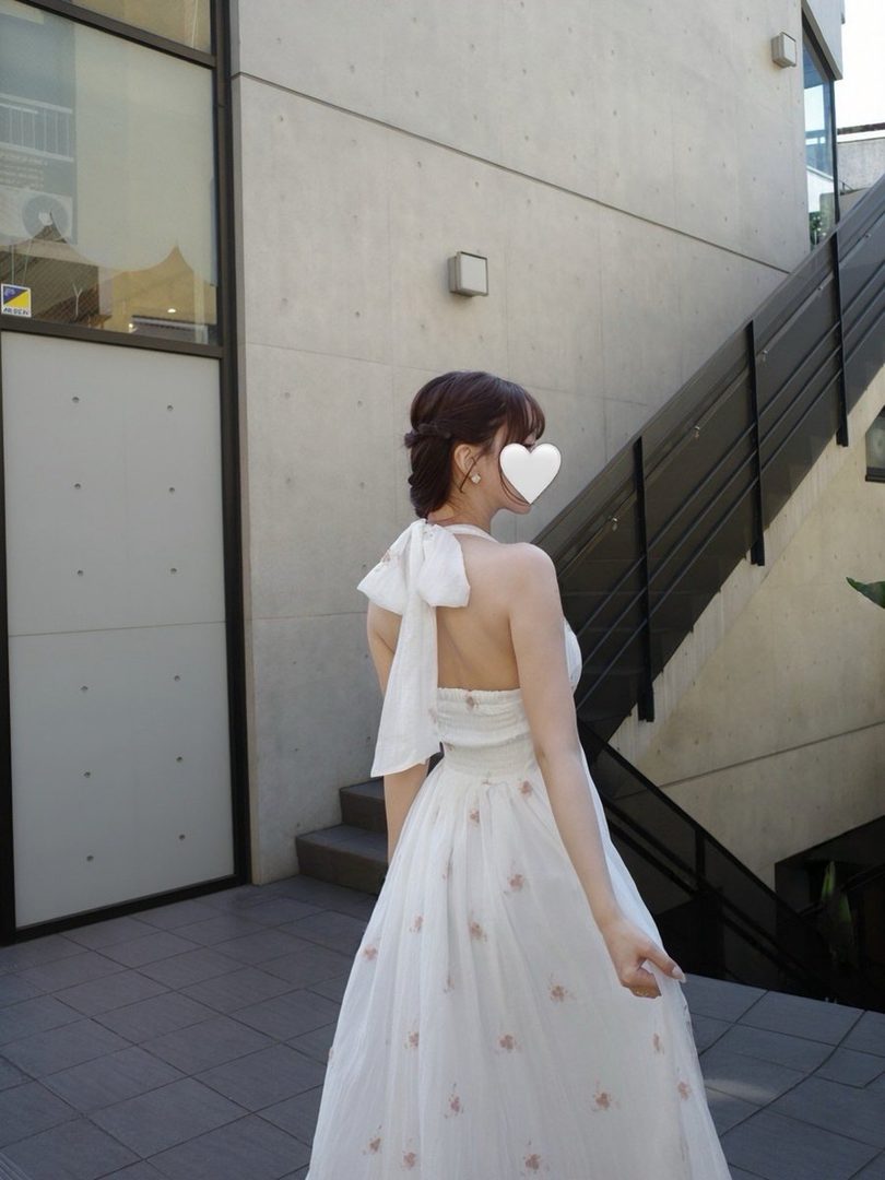 A woman in a white halter dress with floral embroidery stands outdoors, her back to the camera, with a large bow on the back of the dress.