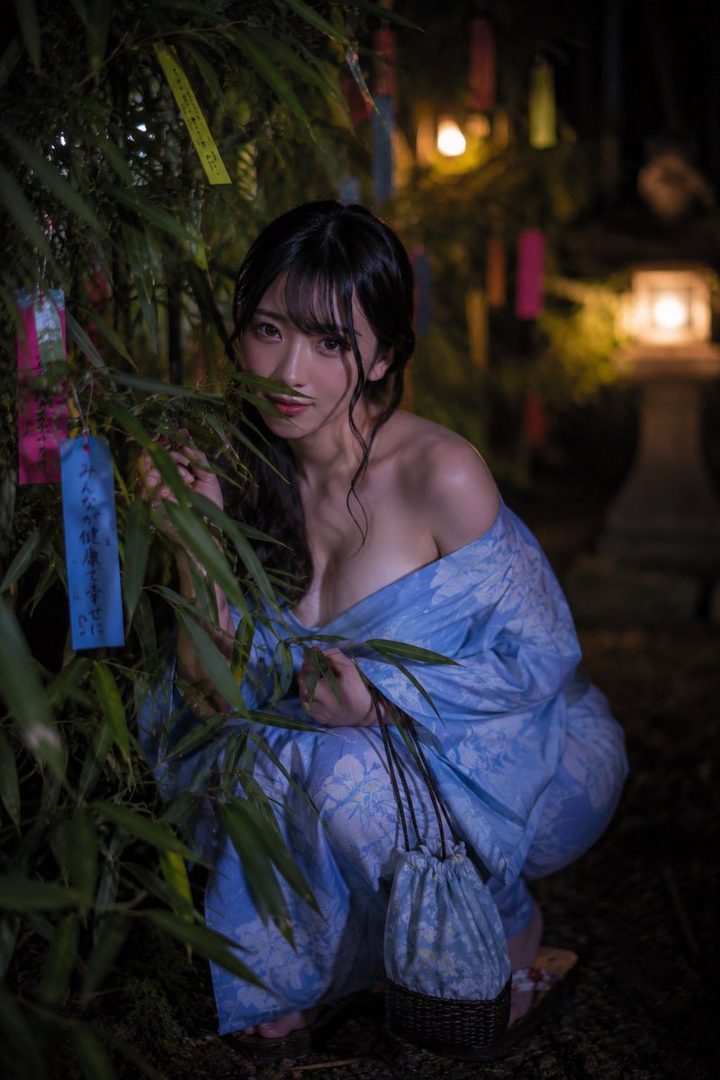 A young woman in a blue yukata crouches near bamboo decorated with colorful tanzaku for Tanabata, looking at the camera with a gentle smile.
