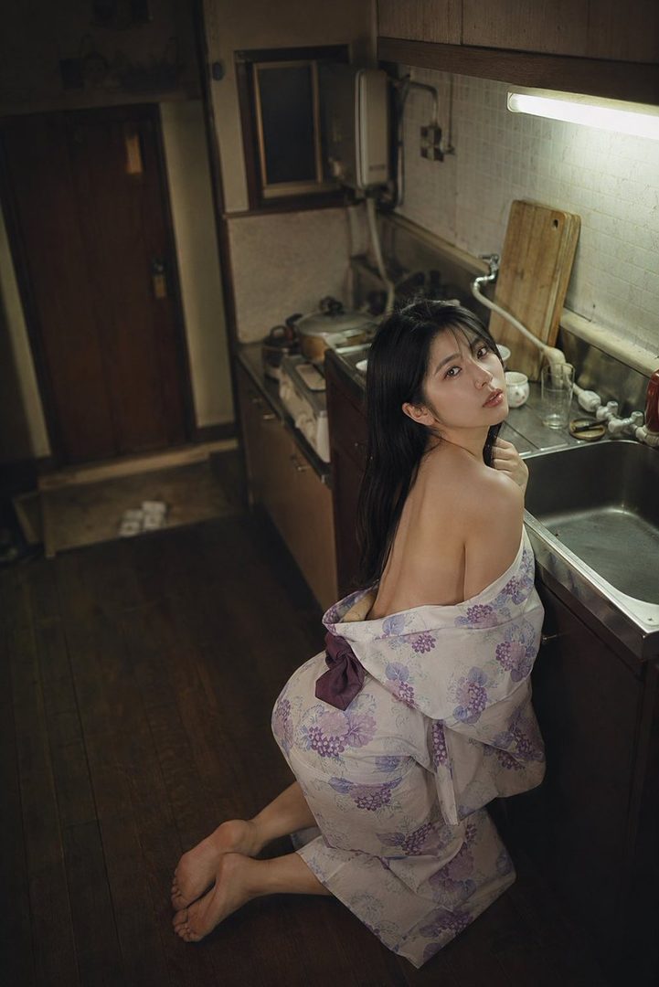 A young woman in a yukata kneels in a dimly lit kitchen, looking over her shoulder.