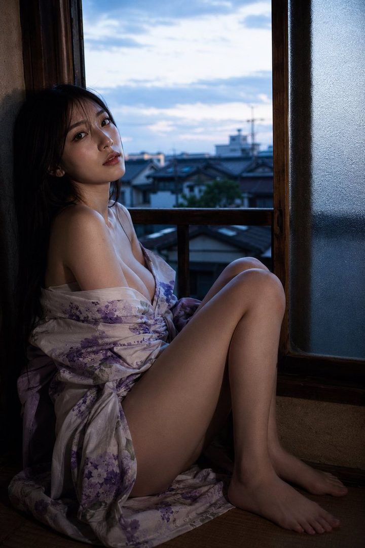 A beautiful East Asian woman with long black hair, wearing a floral robe, poses indoors by a window at dusk.