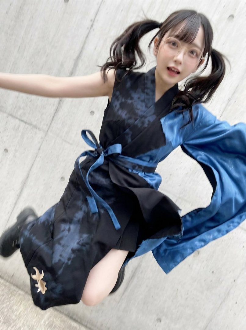 A young woman with twin ponytails and glasses jumps in the air, wearing a blue and black tie-dye kimono-style outfit.