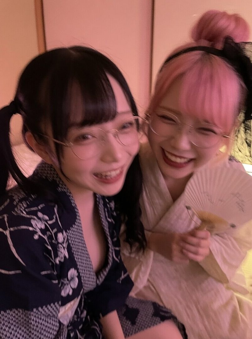 Two young Japanese women in yukatas, one with black pigtails and glasses, the other with pink hair and glasses, smiling and holding a fan.