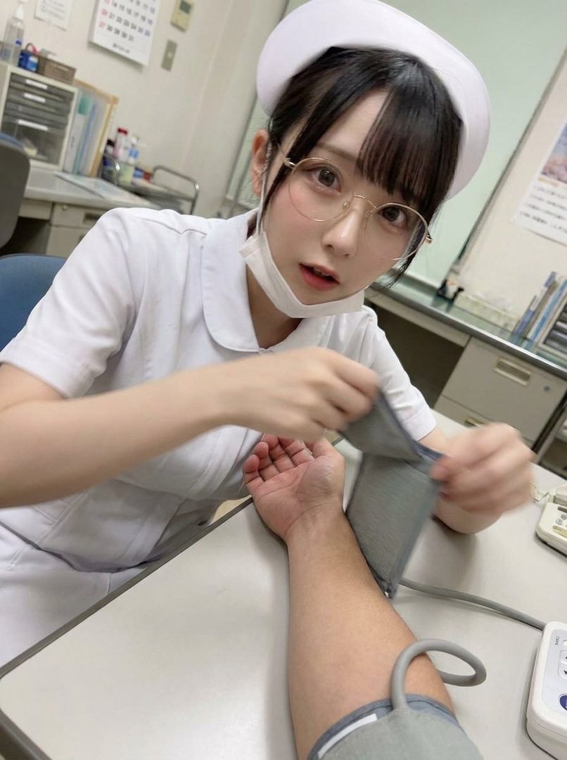 A young nurse in a white uniform and cap, wearing glasses and a mask, takes a patient's blood pressure in a clinic.