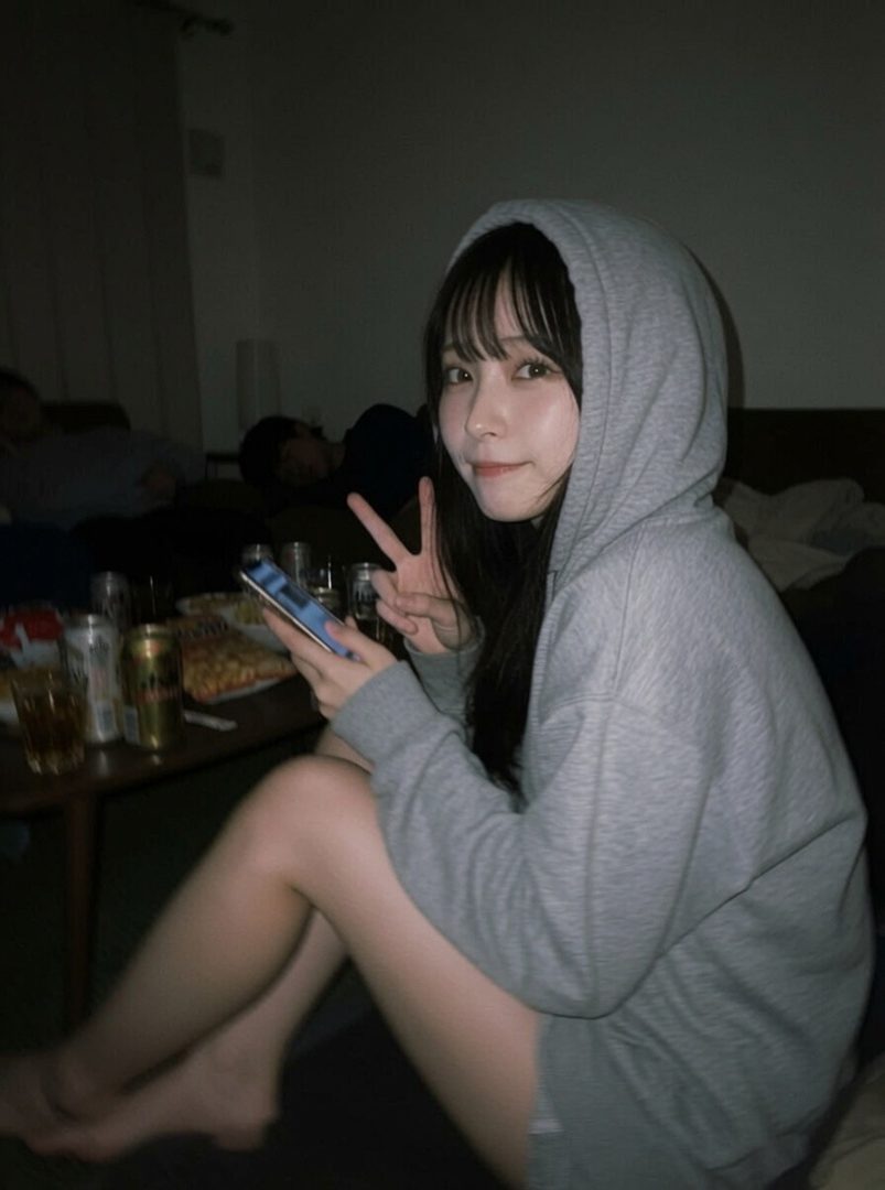 A young woman with long black hair and bangs, wearing a grey hoodie, holds a smartphone and makes a peace sign while sitting indoors.