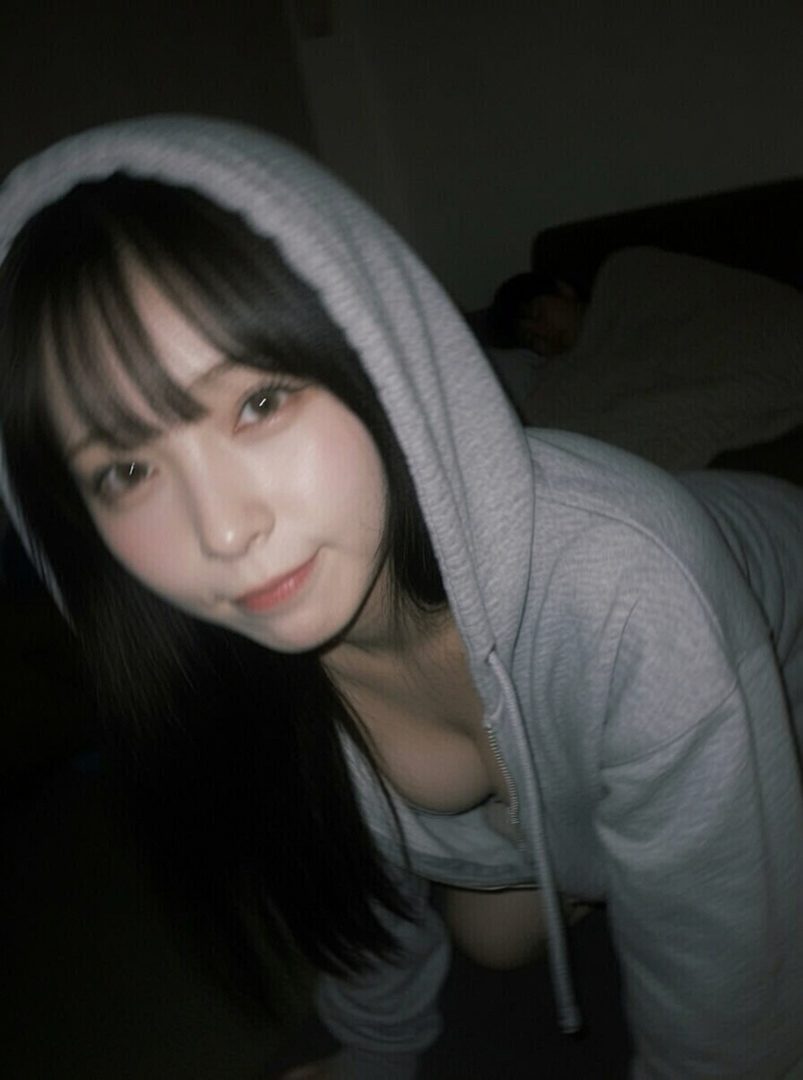 A young woman with long black hair and bangs, wearing a grey hoodie, poses suggestively indoors.