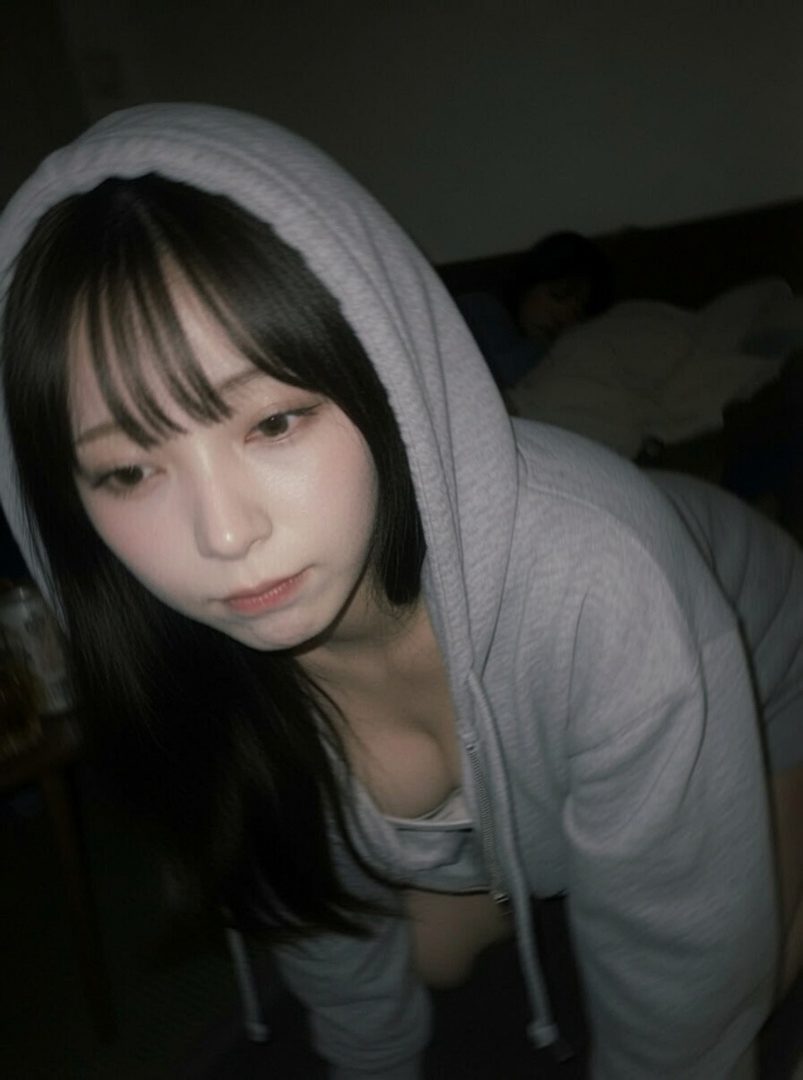 A young East Asian woman with long black hair and bangs, wearing a grey hoodie, poses suggestively indoors.