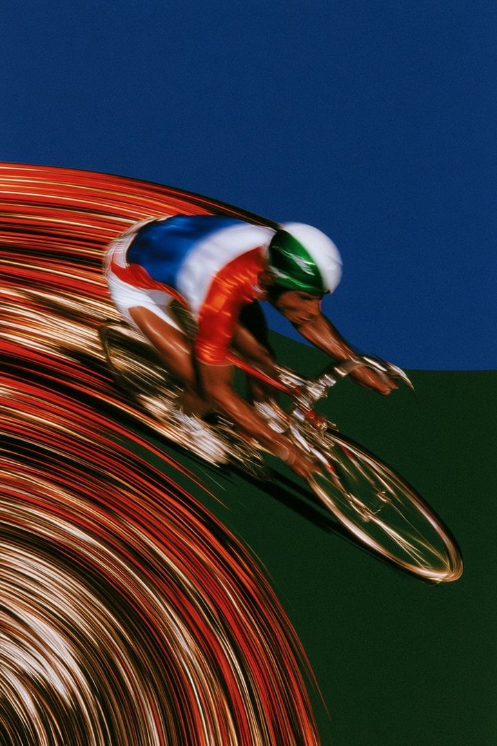 A cyclist in a red, white, and blue racing suit and green helmet speeds down a track, with streaks of light indicating motion.