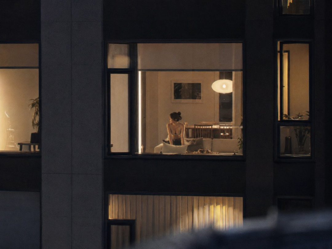 A woman seen through a window at night, wearing lingerie, looking away from the camera.