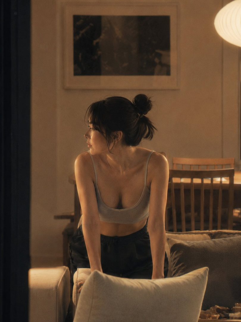 A young woman with her hair in a bun looks out a window in a dimly lit room, wearing a casual top and shorts.