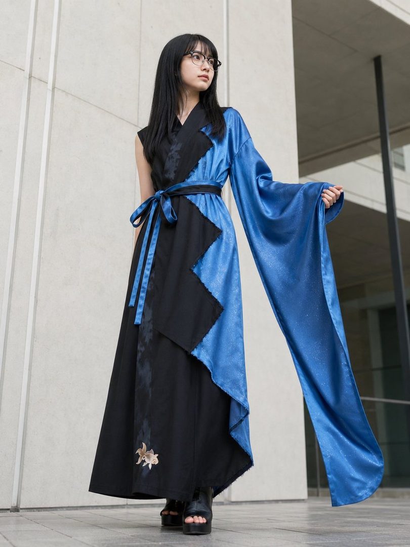 A young woman with long black hair and glasses, wearing a striking blue and black asymmetrical dress with a flowing sleeve, stands outdoors.