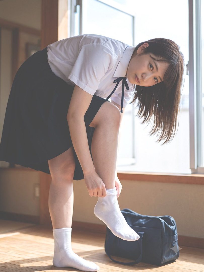 A young woman in a school uniform bends over to adjust her white socks indoors, with natural light coming from a window.