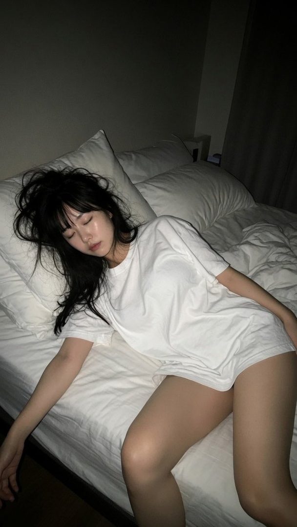 A young woman with long black hair sleeps on a bed in a white t-shirt and shorts, appearing to be drunk.