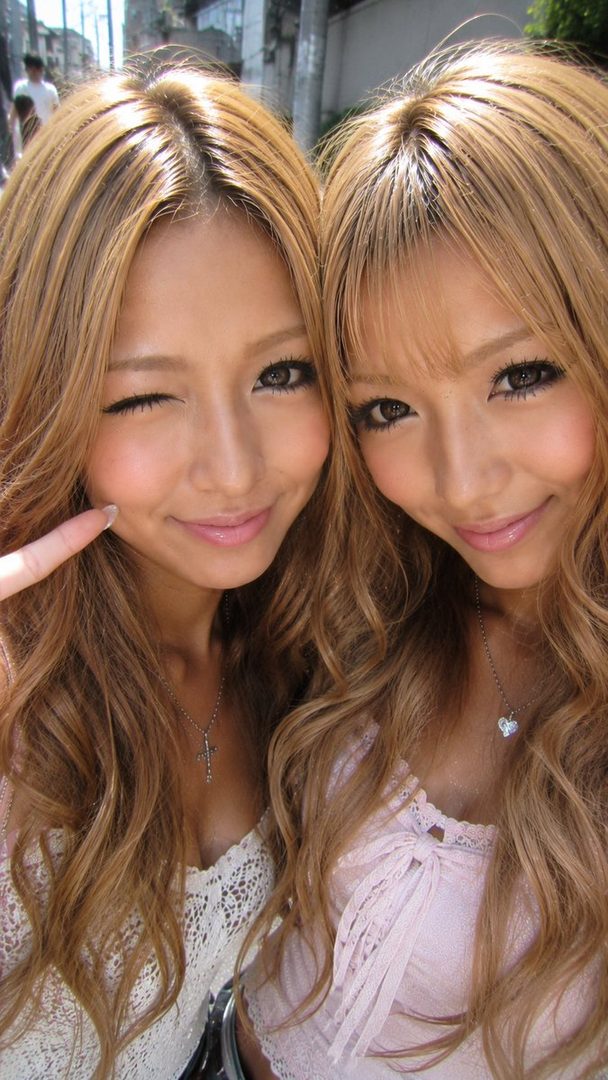 Two young Japanese women with long blonde wavy hair and heavy makeup posing for a selfie outdoors.