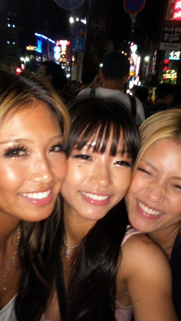 Three young East Asian women smiling and posing for a selfie at night in a city with neon lights.
