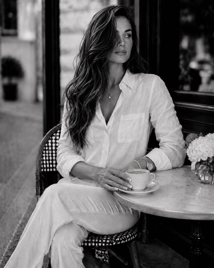 A woman with long, wavy brown hair sits at a cafe table, holding a cup of coffee and looking away from the camera.
