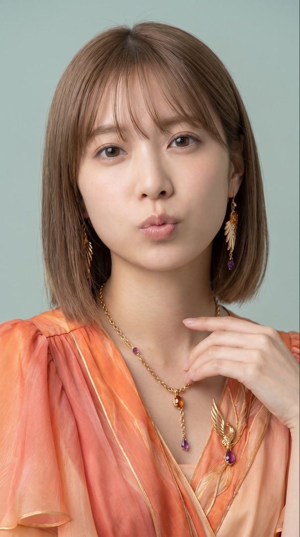 A young East Asian woman with short brown hair and bangs, wearing a flowing orange blouse and elegant jewelry, poses with her lips puckered.