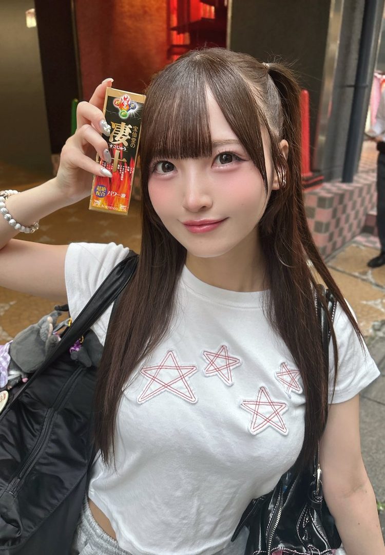 A young East Asian woman with brown hair in pigtails and bangs, wearing a white t-shirt with red star patches, holds up a snack box while smiling at the camera in a street setting.