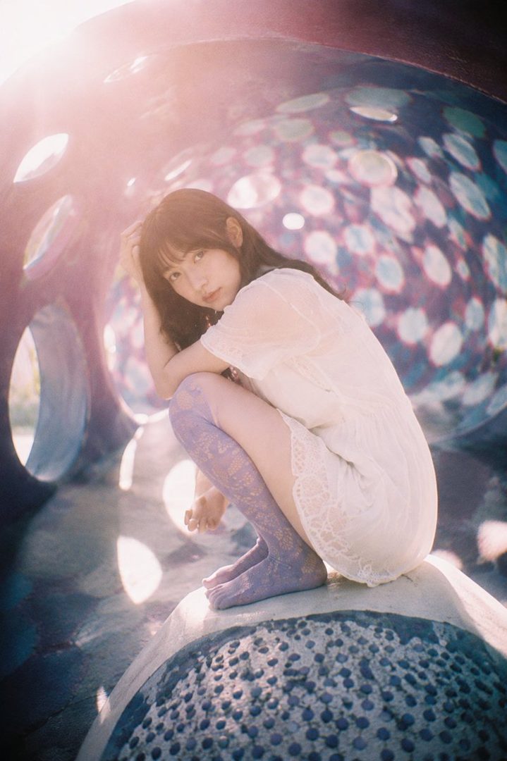 A young woman with long dark hair, wearing a white lace dress and purple patterned stockings, crouches in a colorful, abstract outdoor play structure.
