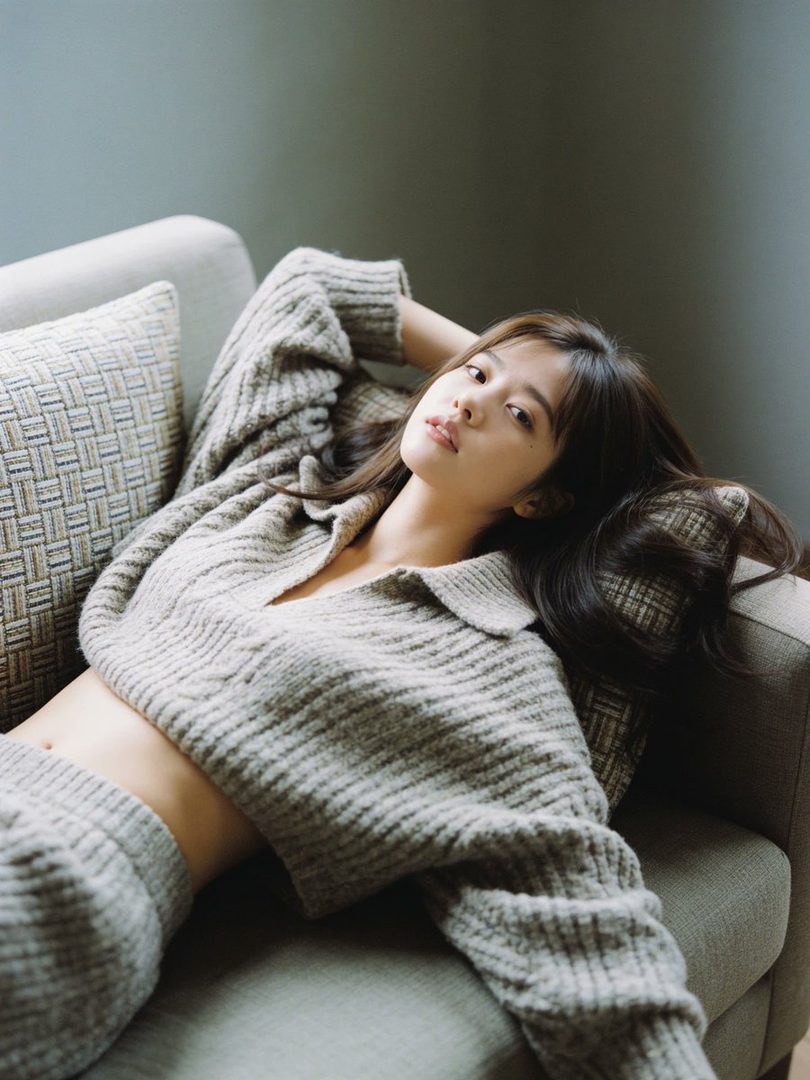 A young woman with long dark hair lies on a sofa, wearing a cozy knit sweater and shorts, with her hand behind her head.