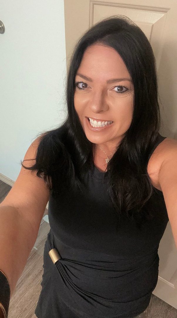 A woman with long dark hair smiles brightly at the camera in a selfie taken indoors.