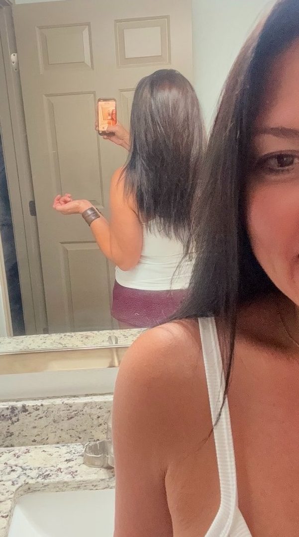 A woman with long, dark hair takes a selfie in a bathroom mirror, wearing a white tank top and burgundy shorts.