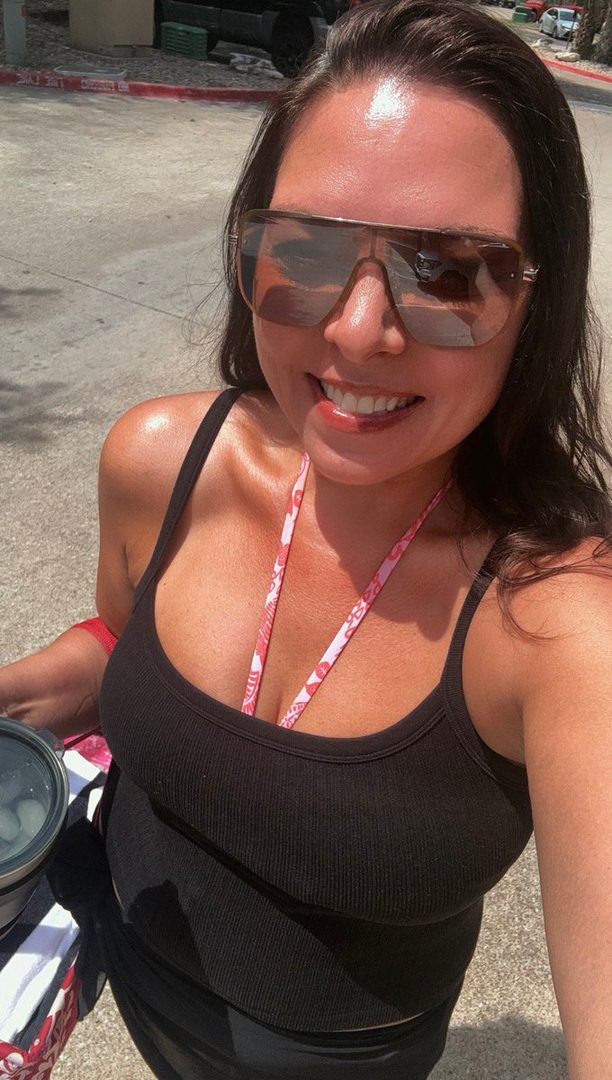 A smiling woman with dark hair and sunglasses takes a selfie outdoors, wearing a black tank top.