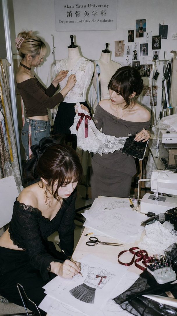 Three young women in a fashion design studio, one sketching a dress, another working on a mannequin, and a third holding fabric.
