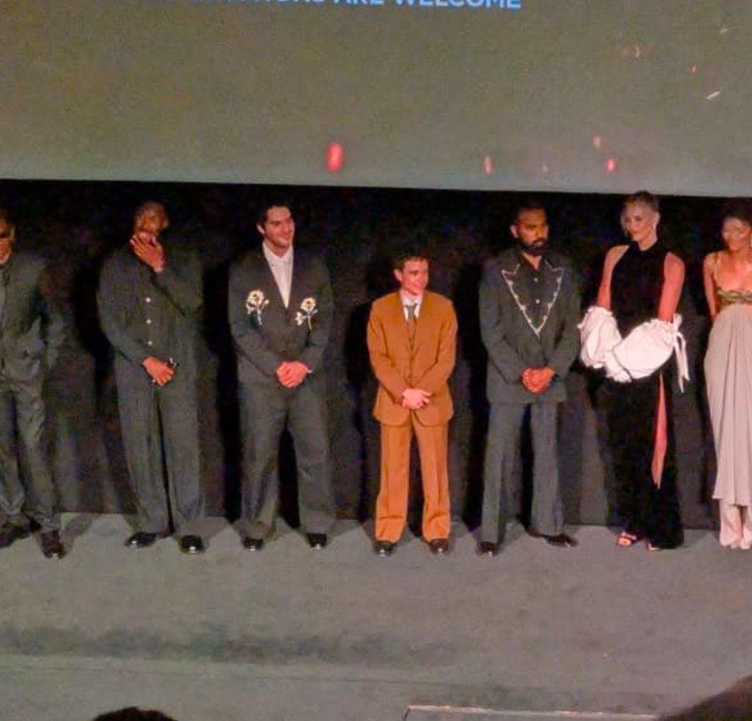 A group of seven people, likely actors or models, stand on a stage in front of a screen, posing for a photograph.