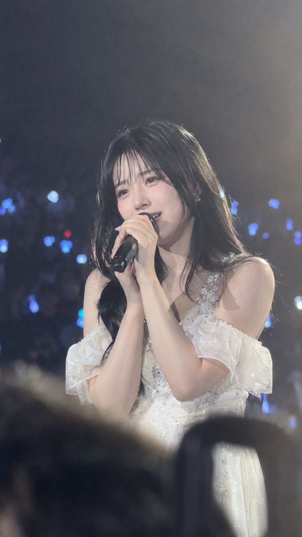 A young East Asian woman in a white off-shoulder gown sings passionately on stage while holding a microphone.
