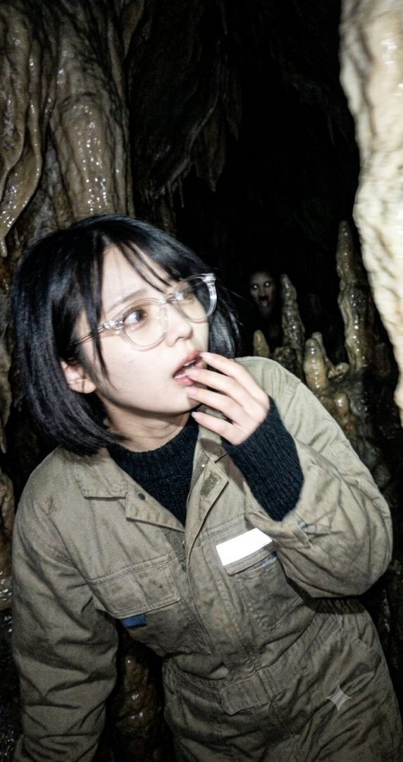 Young woman with short black hair and glasses in a dark cave wearing a jumpsuit, with a ghostly face visible in the background.