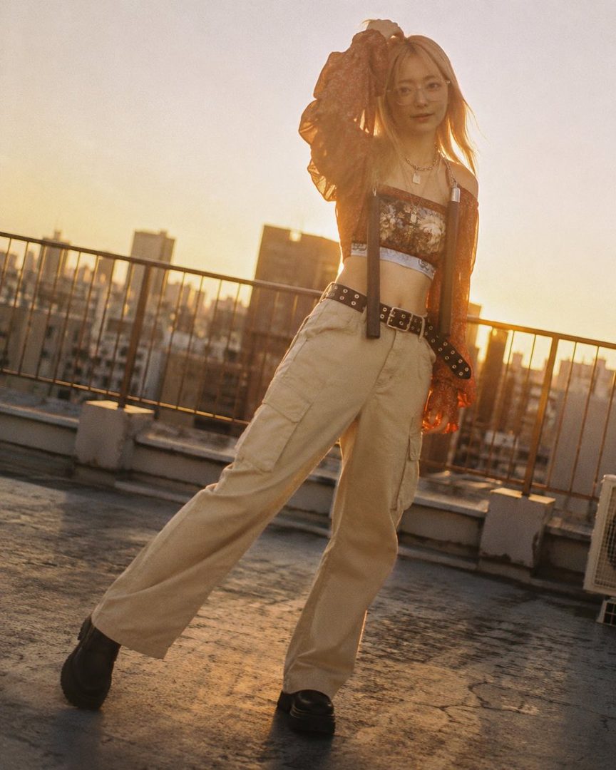 Young blonde woman in stylish crop top and cargo pants posing on an urban rooftop at golden hour sunset.