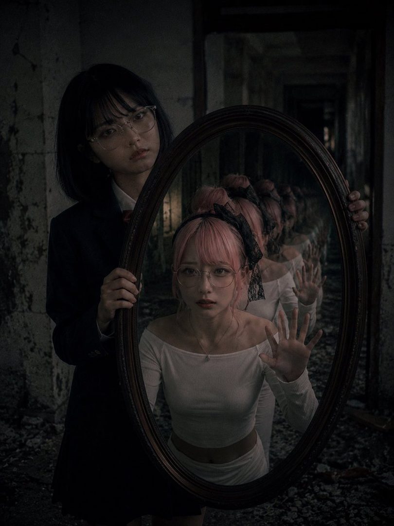 Two young women in an abandoned building, one in school uniform holding a mirror that reflects the pink-haired girl with infinite duplicates in a dark corridor.