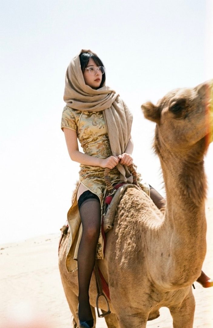 A young East Asian woman in a golden qipao and headscarf rides a camel across a sunlit desert.